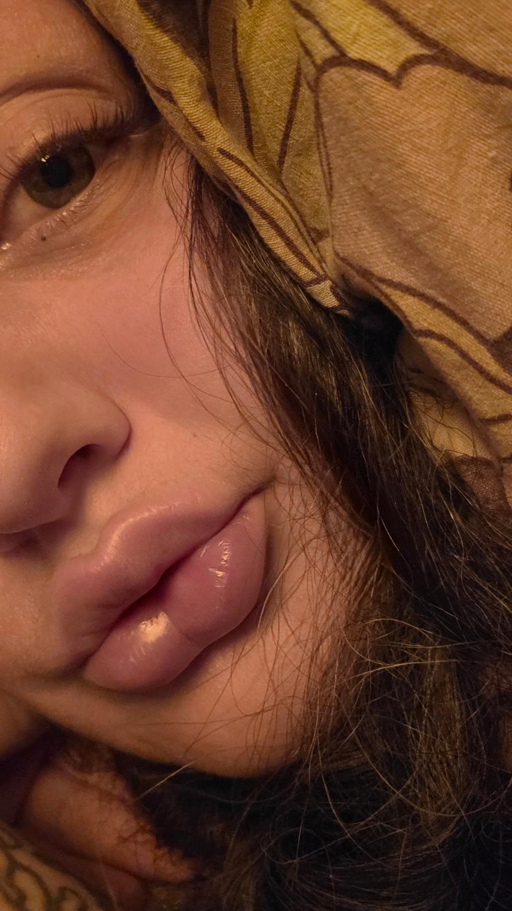 Close-up of a person's face with dark hair and a yellow patterned headscarf or hood, photographed in warm, dim lighting wearing GLOW lashes