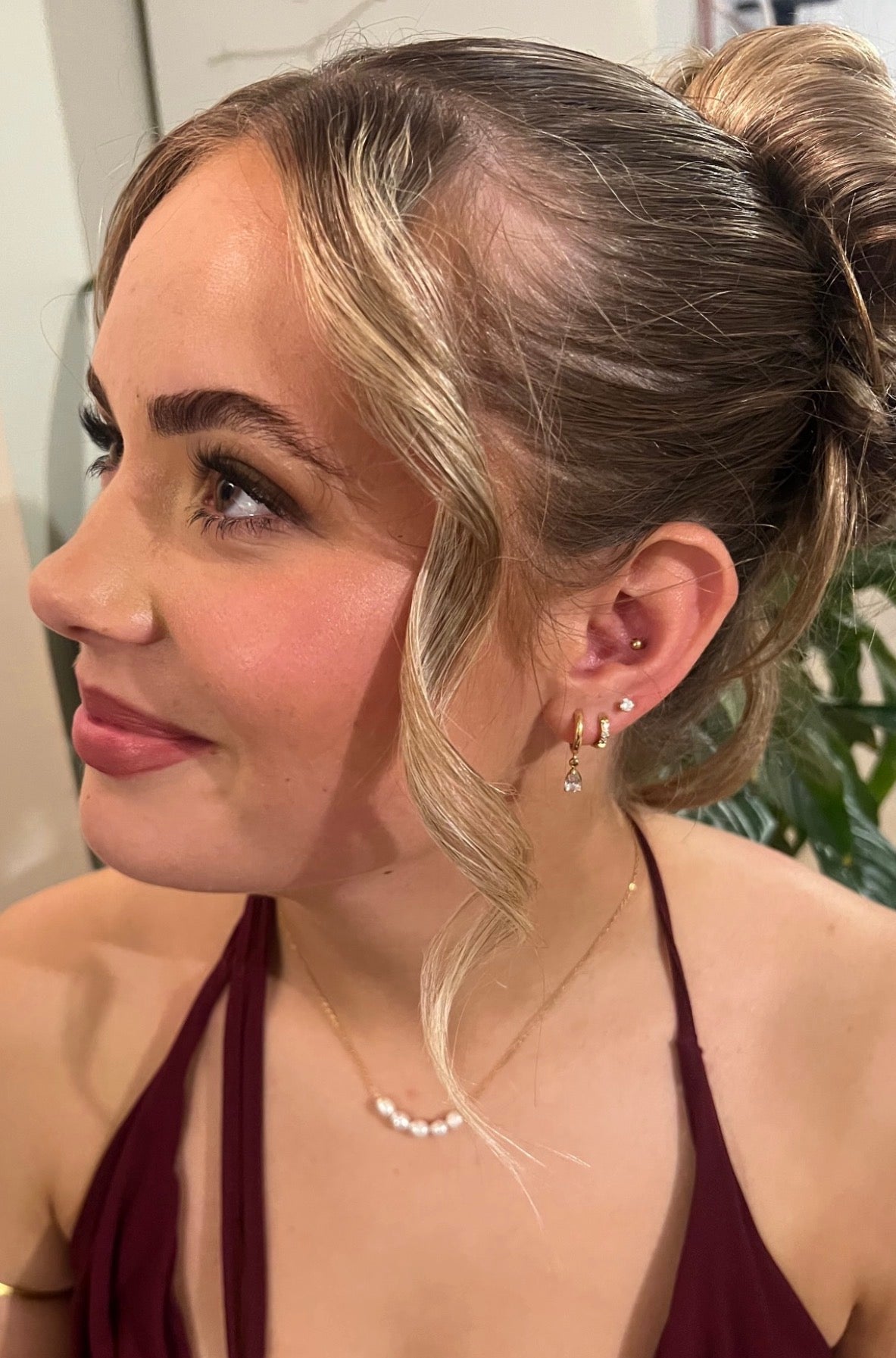 A close-up side profile of a woman with blonde-highlighted hair in a bun, wearing small gold hoop earrings with clear stones and a delicate gold necklace with white pearls, against an indoor background, wearing rove lashes